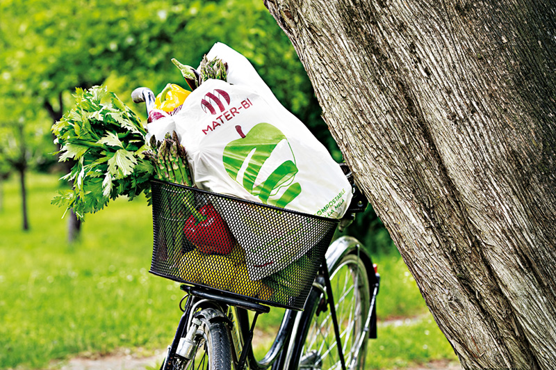Sacchetto in Mater-Bi col disegno di una mela stilizzata, inserito nel cestello portaoggetti di una bicicletta vicino a un albero in un parco